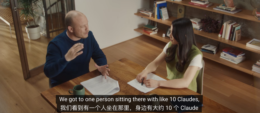 画像は、男性と女性がテーブルを挟んで座り、会話している場面を示しています。男性は濃紺のセーターを、女性は緑色のベストを着用しており、テーブルの上には紙とペンが置かれています。背景には本棚や植物などが見えます。画面下部には中英字幕があり、「私たちは、そこにひとり座っていて、その周りにおよそ10個の Claude がいるのを見ています」といった内容が表示されています。この画像は、AI 支援プログラミングの初期段階に関する説明と関連しており、ひとりの人の前で複数の Claude モデルが補助的に作業する様子を視覚的に示しています。