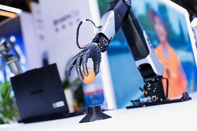 图片展示了BrainCo在WAIC 2026展会上的展示内容,画面中一个机械臂正用黑色手套夹着一个橙色球体。背景左侧有"BrainCo"标识,右侧是穿着橙色上衣的人的图片。该图片与上文提到的BrainCo在WAIC展会上展示的Embodied AI Data Collection Solution相呼应,直观呈现了其研发的机器人在执行复杂物理任务时的场景,体现了其在机器人训练数据收集方面的技术应用。