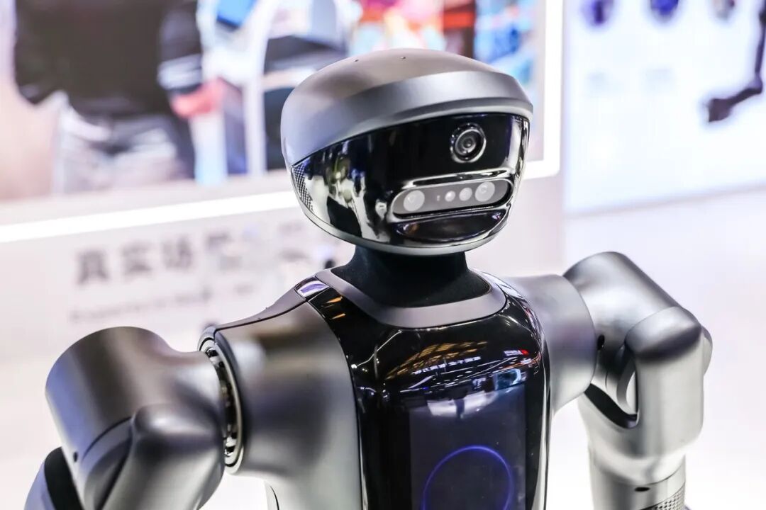 图片展示的是Pudu Robotics在WAIC 2026上展出的工业半人形机器人PUDU D7。其外观为银色金属材质,头部呈圆顶状,带有摄像头,面部有多个传感器,胸前有蓝色圆形区域。背景中可见“真实场景”等字样,以及一些模糊的宣传海报。PUDU D7是Pudu Robotics“一脑多形”战略的硬件体现,与文档中介绍的PuduFM 1.0和PuduAgent等技术相呼应,展示了公司在机器人领域的最新成果。