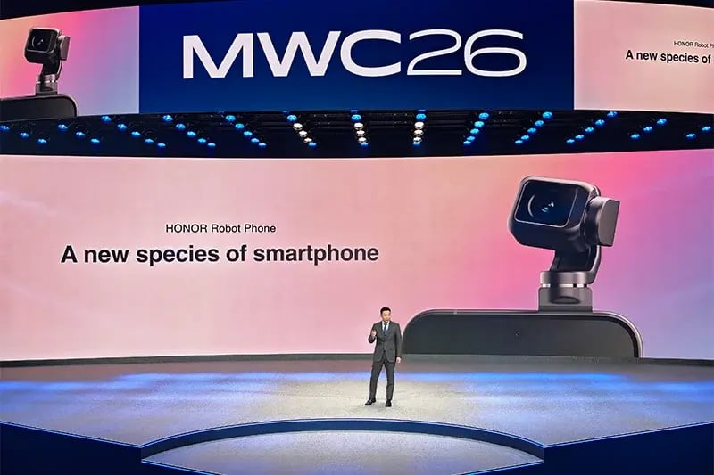 图片展示的是HONOR在MWC26(世界移动通信大会2026)上发布Robot Phone的场景。画面中,一位男士站在讲台上,背景大屏幕上显示“MWC26”及“HONOR Robot Phone A new species of smartphone”字样,右侧还有一台带有镜头的设备。该图片与上下文紧密相关,上下文介绍了HONOR Robot Phone的核心机械特征、自研微电机、相关功能及官方产品宣称,此图直观呈现了发布会现场情况,与产品发布主题相契合。