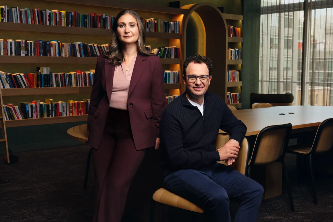 この画像は、Dario Amodei氏とDaniela Amodei氏の二人を表しており、書棚と会議テーブルが配置されたオフィス空間にいる様子である。Daniela Amodei氏はワインレッドのスーツに淡いピンクのインナーを合わせ、やや左寄りに立っている。Dario Amodei氏は濃い色のニットカーディガンと濃紺のパンツを着用し、椅子に座っている。二人の表情は穏やかである。背景の書棚にはさまざまな書籍が並び、右側の会議エリアにはテーブルと椅子が配置されており、文書で言及されている二人がAnthropicの共同創業者であるという設定に合致し、二人の仕事に関連する姿を視覚的に示している。